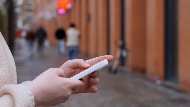 Close-up hands hold smartphone while walking down urban alley. Background shows blurred pedestrians and brick walls. Person appears focused, absorbed in device. Suitable for themes like technology