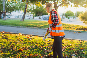 Obraz premium Woman wearing a safety vest and gloves, raking fallen autumn leaves in a sunny park lawn, undertaking community service as a volunteer for outdoor cleaning and park maintenance