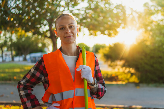 Female street sweeper wearing a high visibility vest and gloves, holding a broom, smiling at the camera, working during sunset in an autumn park, contributing to public sanitation