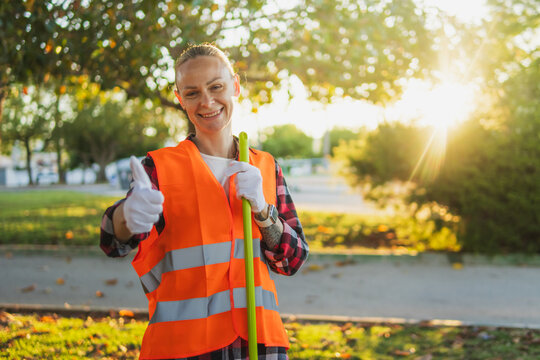 Smiling woman in orange safety vest and gloves gives thumbs up while holding a garbage picker during a sunny community cleanup, promoting environmental stewardship and urban care