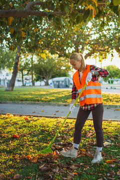 Woman in a reflective vest and gloves raking dry leaves from the grass in a sunny park, actively participating in outdoor maintenance and community service