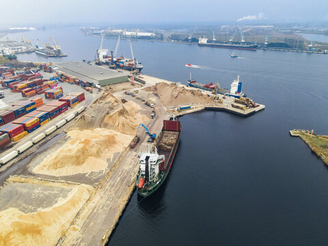 Riga, Latvia - 03 November 2021: Aerial view of the busy commercial port with cargo ships, shipping containers, and piles of wood chips being loaded for transport.