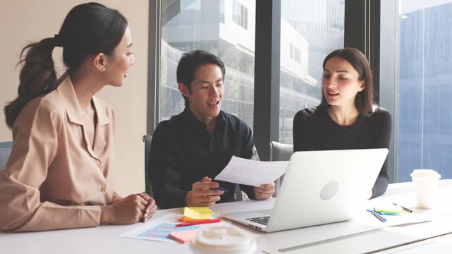 Three business professionals discussing charts and reports while analyzing information on laptop during collaborative office meeting.