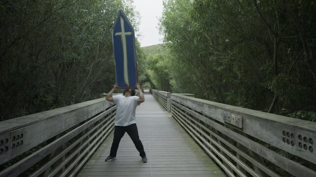 Man Balancing Blue Cross Sign on Head on Wooden Boardwalk - Wide, Steady Cam Shot