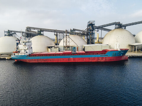 Riga, Latvia - 03 November 2021: Aerial view of a cargo ship docked at the port of Riga, featuring large industrial storage domes and conveyor systems in the background.