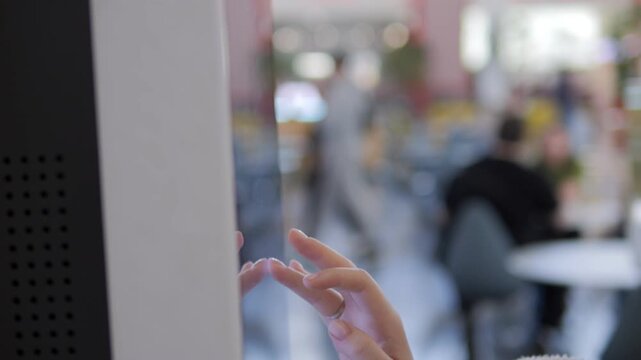 Child's hand gently touches a modern touchscreen kiosk. The background shows a softly blurred cafe or public space with people seated at tables. The focus remains on the child's curiosity