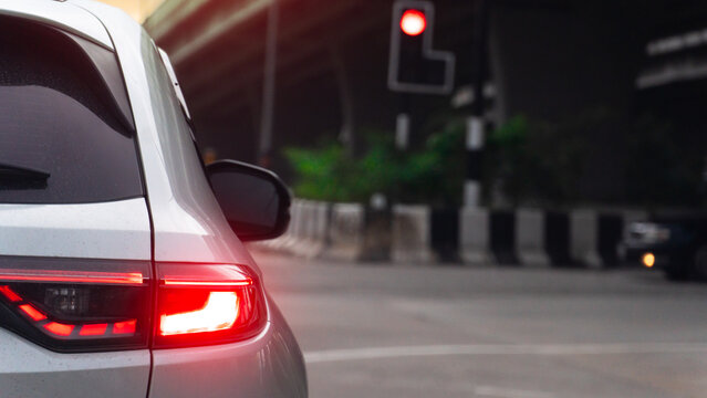 Tight rearview shot focusing on the bright red tail light. White car waiting at an urban road intersection during dusk. Red traffic light glows softly and intersection under the overpass