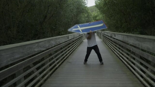 Man Spinning Painted Board On Wooden Footbridge Among Trees - Steady Cam Shot