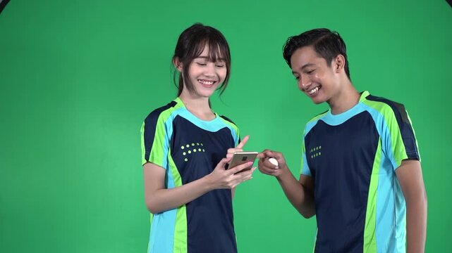 Near-end frame shows young woman and man in matching sportswear, smiling, talking, and giving thumbs up while checking smartphone against bright green studio