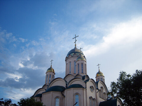 Low-angle shot of majestic orthodox church with golden and green domes topped with crosses. The brick cathedral building stands against a bright blue summer sky filled with dramatic white clouds