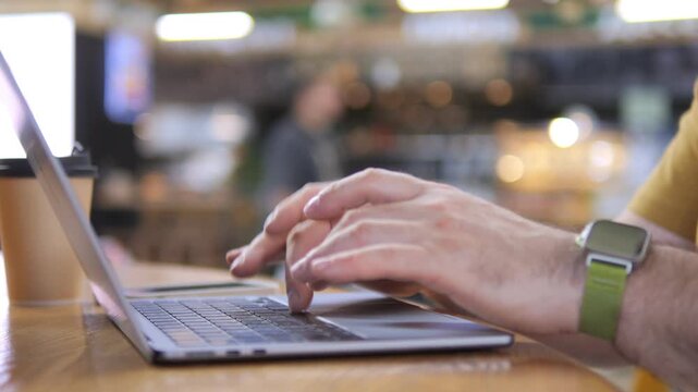 Hands type on laptop in cozy cafe. Person wears smartwatch and yellow shirt. Focus on keyboard and fingers. Background softly blurred with cafe ambiance. Ideal for remote work, digital lifestyle
