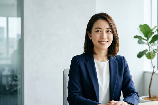 オフィスでカメラ目線で笑顔で座る紺色ジャケットの30代日本人女性