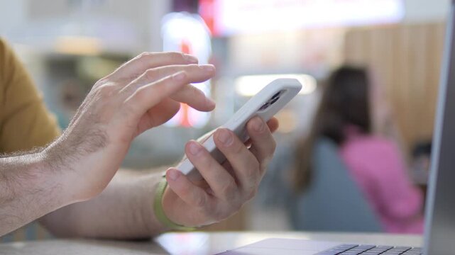 Hand scrolls smartphone at cafe table. Background shows blurred person in pink. Scene suggests casual digital activity. Can use for tech, communication, or work-from-cafe themes
