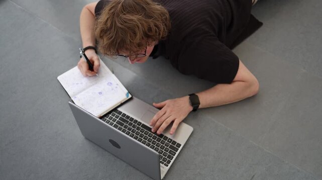 Student studies while lying on floor using notebook and laptop showing dedication to education determination continuous learning academic effort and focus