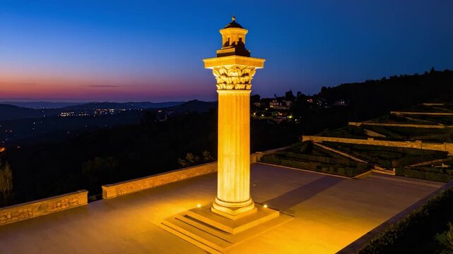 A tall, illuminated classical column on a patio overlooking a hillside landscape at dusk