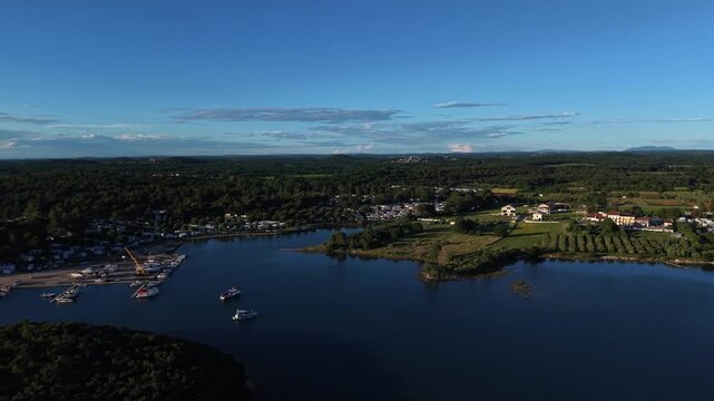 Marina filled with boats sits along forested peninsula in Istria, Croatia, with tranquil bay separating coastal neighborhood from green fields beneath clear evening sky. Aerial drone approaching