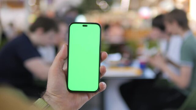 Hand holds phone with green screen. Blurred cafe background shows people seated. Focus remains on device, ideal for app or social media mockups. Casual, relaxed atmosphere with soft lighting
