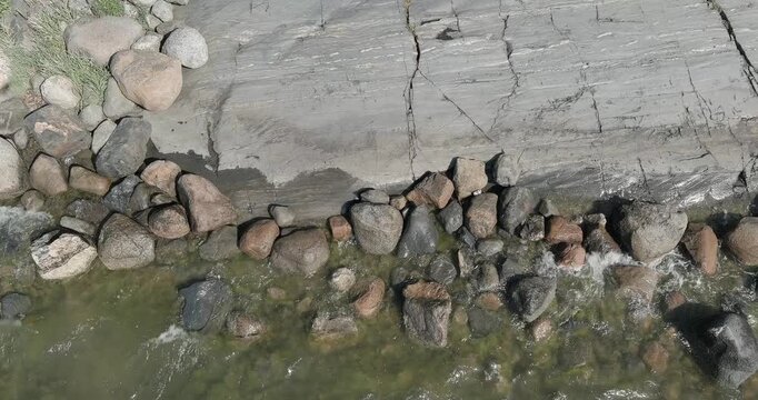 Rocky shoreline or littoral zone, where a massive slab of bedrock meets loose, rounded cobbles and stones in shallow water in summer, aerial view, Uutela, Helsinki, Finland, Europe.