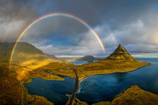 Aerial view of the iconic Kirkjufell mountain with a vibrant double rainbow arching over the surrounding coastal landscape and calm waters in Grundarfjordur, Iceland.