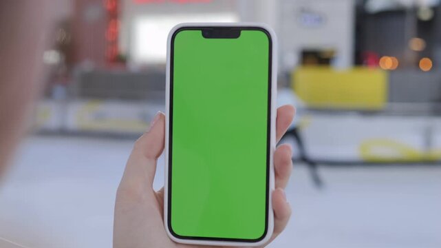 Hand holds phone with green screen. Background shows blurry ice rink. Person focuses on device. Useful for apps, UI mockups, or advertising. Realistic setting with soft focus