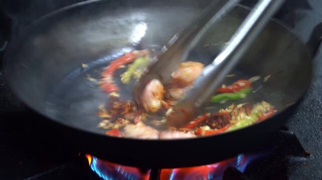 Chef stirring spicy shrimp and vegetables in hot frying pan over blue gas stove flames.