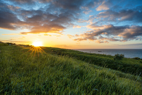 Beautiful sunset over the coastal hills of Arromanches-les-Bains in Normandy. Golden sun rays shining through green grass field overlooking the sea and WWII historic D-Day location in France.