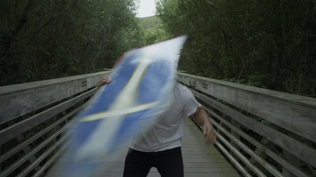 Man Spinning Painted Board On His Head on Wooden Footbridge - Medium Shot