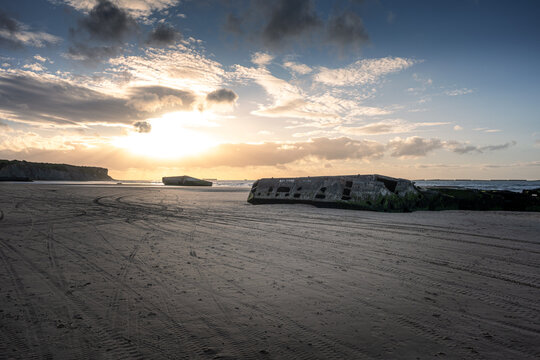 Remnants of the Mulberry artificial harbour on the sandy beach of Arromanches-les-Bains in Normandy. Historic WWII D-Day landing site at Gold Beach during sunset with dramatic sky in France.