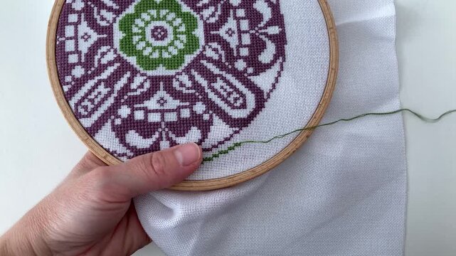 A woman embroiders a purple-green floral ornament, traditional cross-stitch. Close-up of a woman's hands with a wooden hoop, an art of needlework
