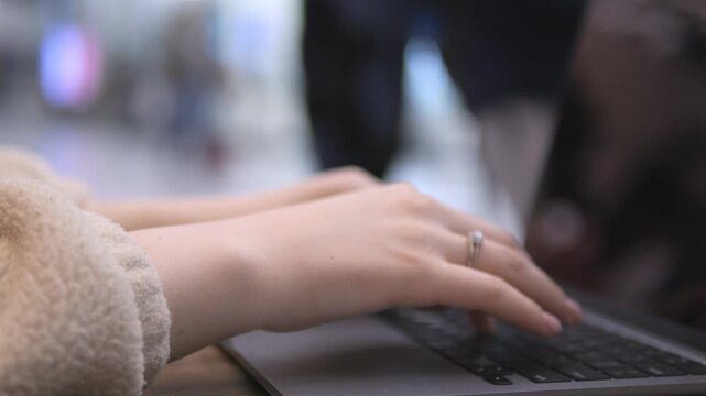 Close-up of hands typing on laptop keyboard. Worn cozy sweater sleeve visible. Ring on finger. Background blurred with soft bokeh effect. Hands move steadily across keys