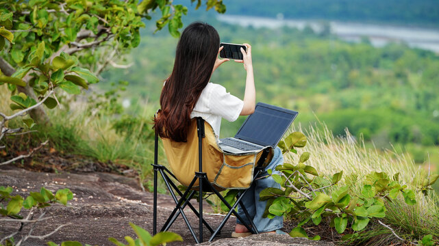 Young woman working remotely on laptop while taking smartphone photo in nature