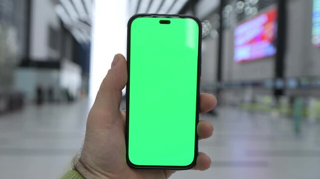 Hand holds phone with green screen. Airport terminal background blurred. Person uses device for travel. Green screen ready for overlay. Modern building with digital displays visible.
