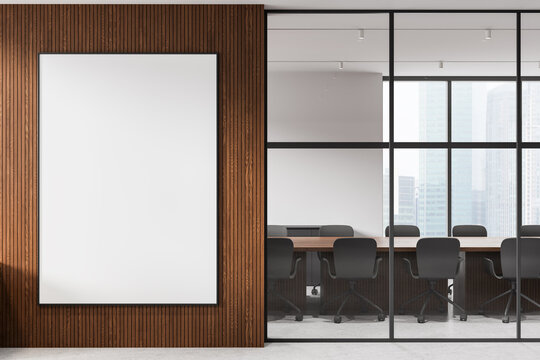 Modern interior with large blank vertical frame on wooden wall beside glass-walled meeting room featuring chairs, table and city view in bright office space. 3D Rendering