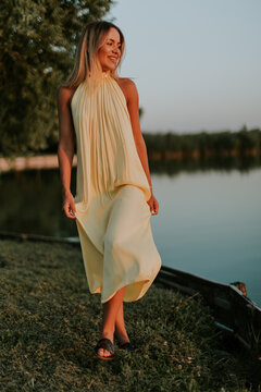 Woman walks by lake wearing yellow dress at sunset in summer time