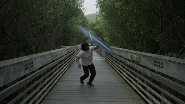 Man Dancing and Spinning Painted Board on Wooden Footbridge - Wide, Steady Cam Shot
