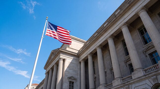 The American flag waving on a tall silver flagpole in front of a grand neoclassical stone building with massive columns under a clear blue sky