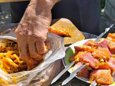 Close-up of hands preparing marinated meat for grilling outdoors. Pieces of raw meat coated with seasoning and marinade are being cut and threaded onto metal skewers. Plastic bag with marinated meat, 