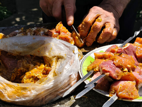 Close-up of hands preparing marinated meat for grilling outdoors. Pieces of raw meat coated with seasoning and marinade are being cut and threaded onto metal skewers. Plastic bag with marinated meat, 