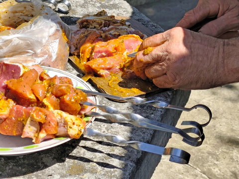Close-up of hands preparing marinated meat for grilling outdoors. Pieces of raw meat coated with seasoning and marinade are being cut and threaded onto metal skewers. Plastic bag with marinated meat, 