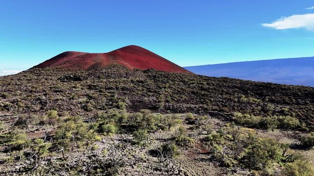 Aerial view of the red cinder cone of Mauna Kea volcano with volcanic landscape and sparse vegetation under a clear blue sky in Hawaii County, Hawaii, United States.