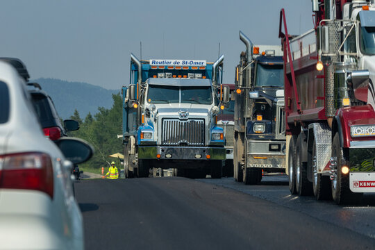 Quebec, Canada, 29 June 2026 : Heavy dump trucks and passenger vehicles navigate a road with a construction worker present.