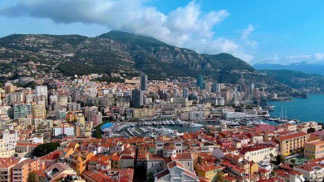 Aerial flight over Monaco Cathedral and the old town of Monaco Ville toward Port Hercules, with superyachts in the marina, Monte Carlo high rise skyline and mountains above the Mediterranean coast.
