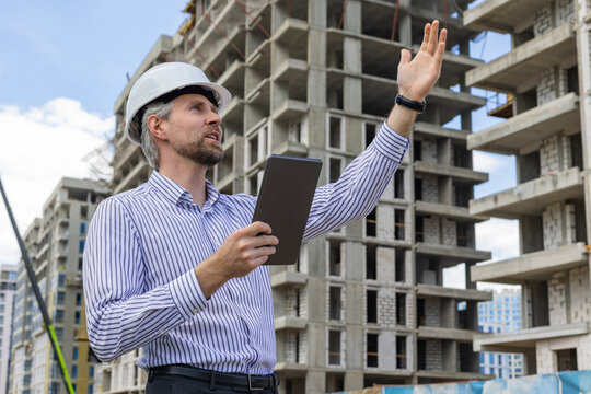 Side view of male engineer standing against background of buildings under construction working in construction helmet