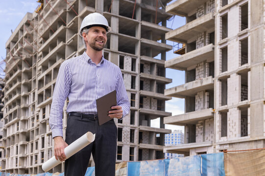 Male engineer standing against background of buildings under construction working in construction helmet