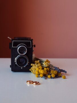 Vintage twin-lens reflex camera, gold wedding rings, and Sardinian wildflowers on a white table with pink wall background