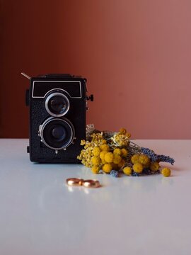 Vintage twin-lens reflex camera, gold wedding rings, and Sardinian wildflowers on a white table with pink wall background