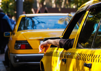 A taxi driver sits in a yellow taxi with their arm resting outside the window. Other taxis fill the background. People walk by on the street. It is a sunny day. © Sergo