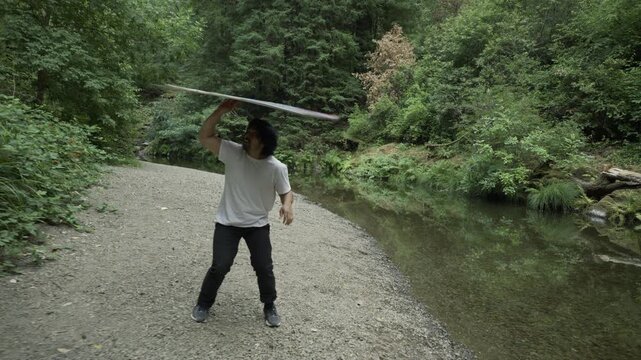 Man Spinning Painted Board Overhead on Gravel Riverbank - Steady Cam Forward
