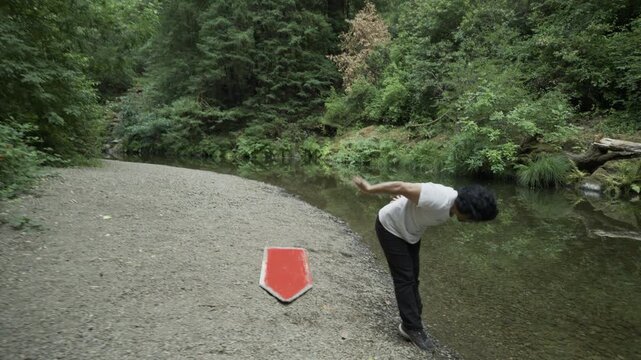 Man Spinning Painted Board And Releasing It Mid-Swing On Gravel Riverbank - Wide Shot