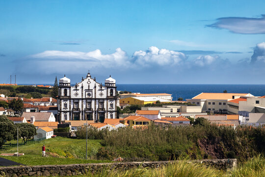 Panor&aacute;mica del pintoresco pueblo de Santa Cruz das Flores en Azores con una imponente iglesia barroca de piedra blanca y negra frente al mar azul, bajo un cielo con nubes esponjosas.